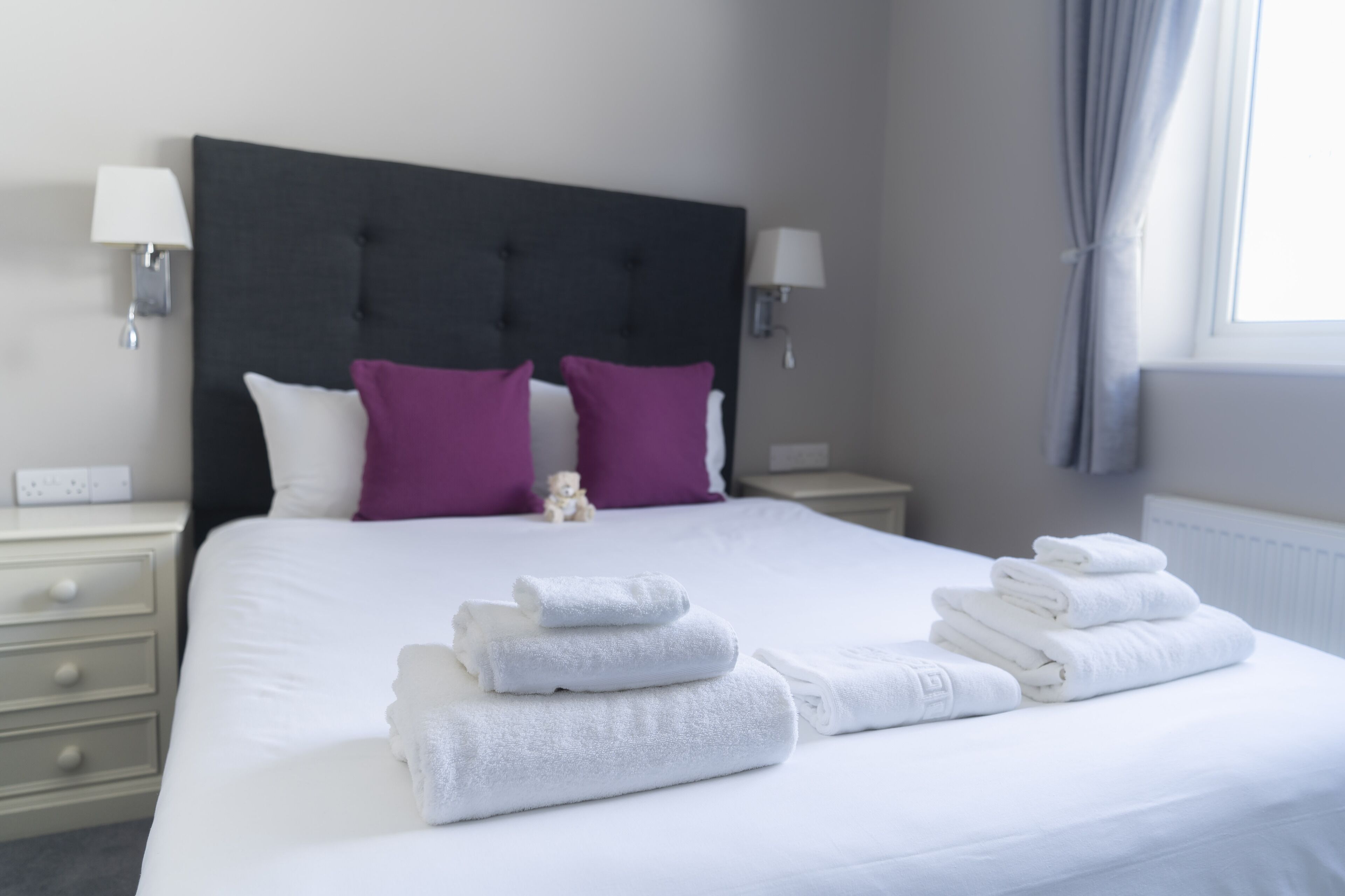 A neatly made bed with white linens and folded towels, a dark headboard, and two purple pillows.
