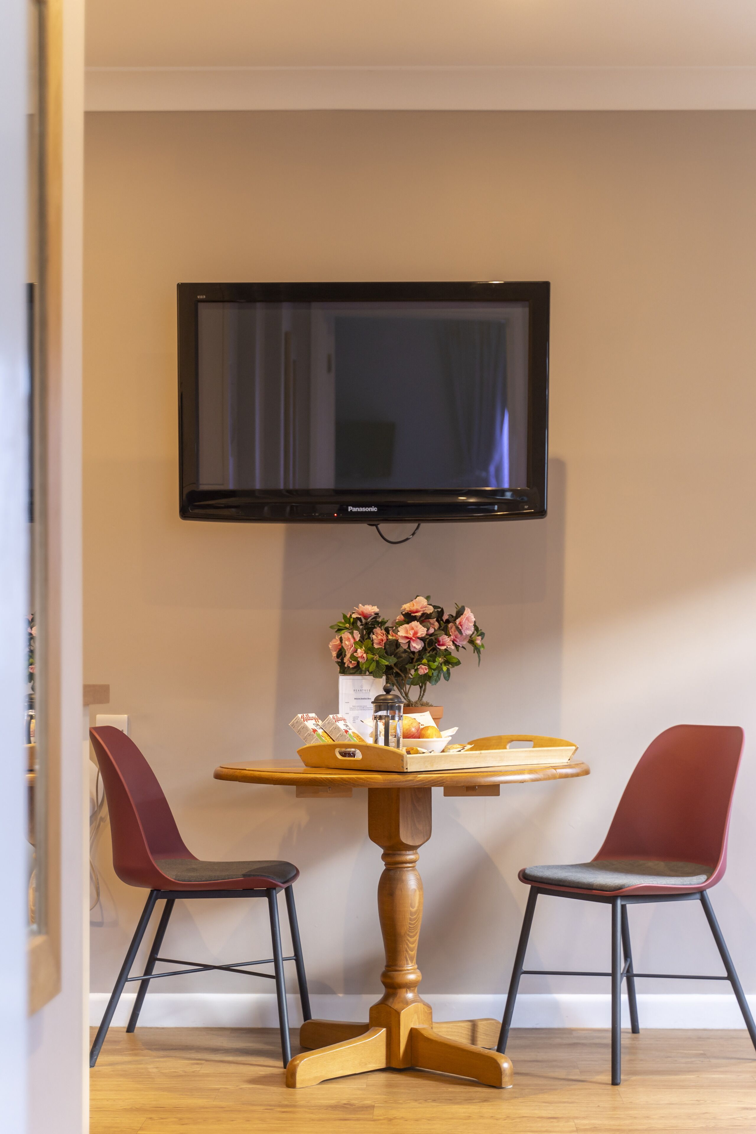 A dining area with a wooden table, two chairs, a television mounted on the wall, and a vase with flowers on the table.