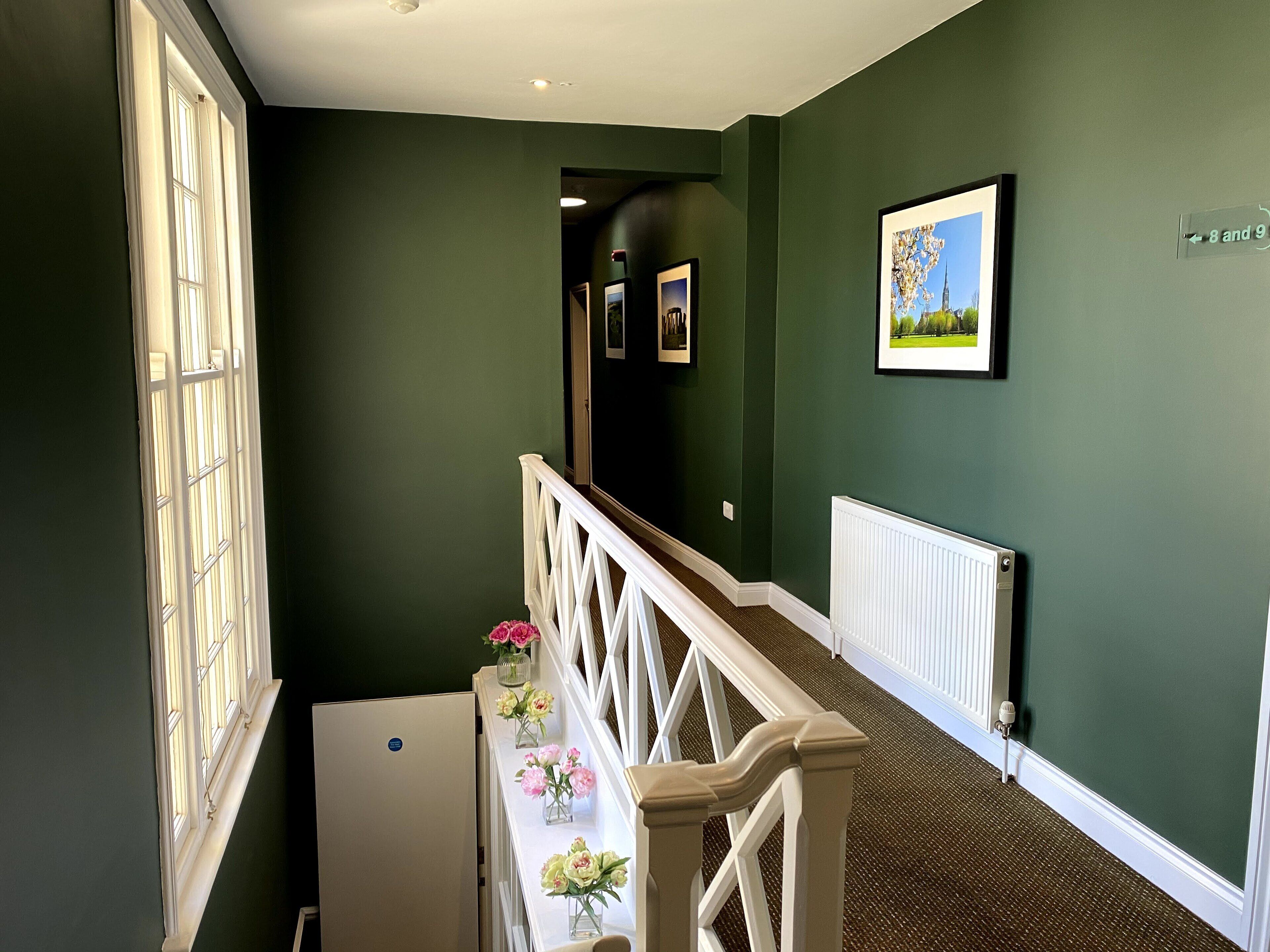 A hallway with green walls, a white railing, and a radiator. There are framed pictures on the wall and a window with blinds.