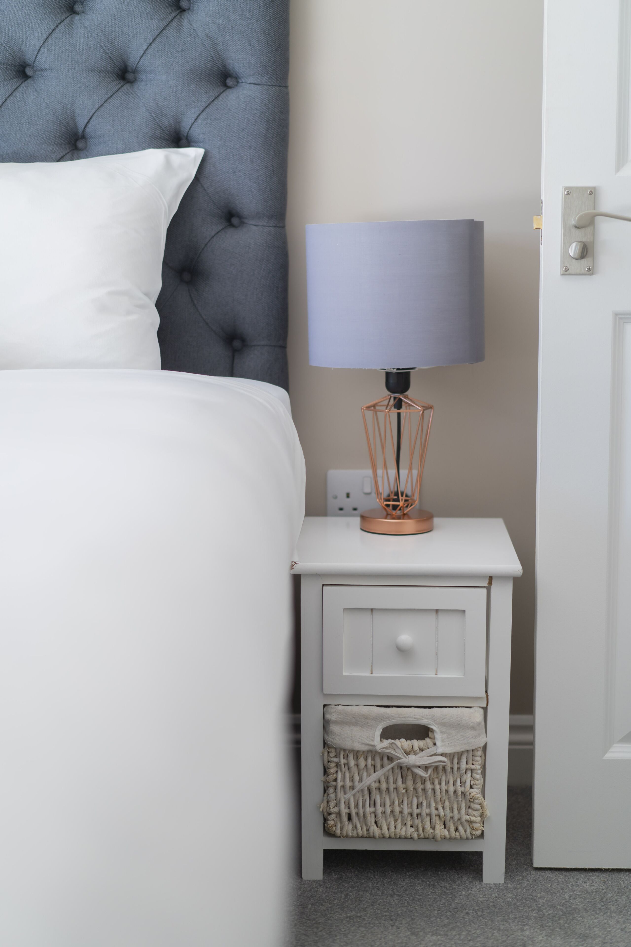 Een netjes opgemaakt bed met een gestoffeerd hoofdbord, een bedlampje en een klein wit nachtkastje met lades en een mandje.