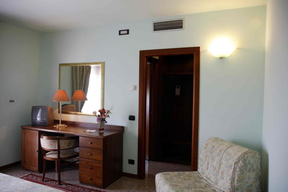 Ein Zimmer mit Schreibtisch aus Holz, einem Stuhl, einem Fernseher, einem Spiegel, einer Lampe und einem sofasartiges Möbelstück mit Blumenmuster.
