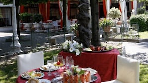 Un área de comedor al aire libre con mesas dispuestas para una comida, rodeada de follaje y detalles arquitectónicos.