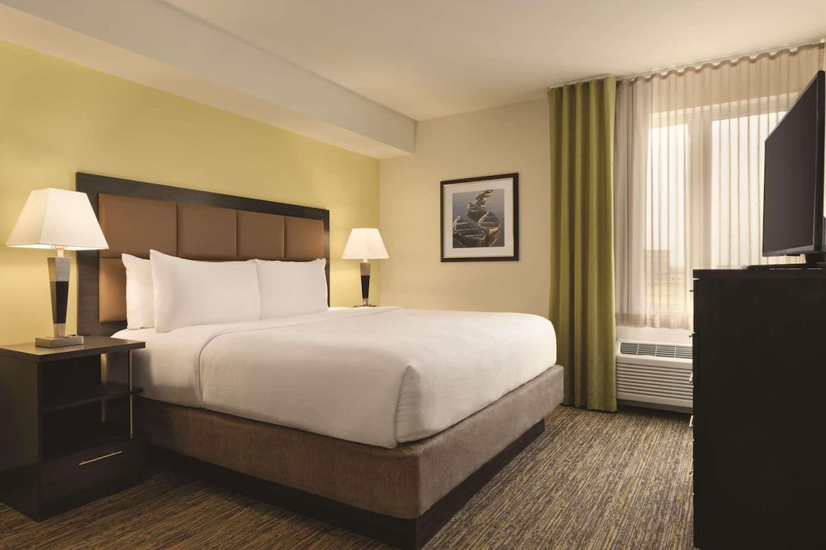 A hotel room with a large bed, two bedside lamps, a flat-screen TV, an air conditioning unit, and a framed picture on the wall.