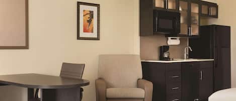 A hotel room with a bed, a desk, a chair, a kitchenette with a microwave, a refrigerator, and a framed picture on the wall.