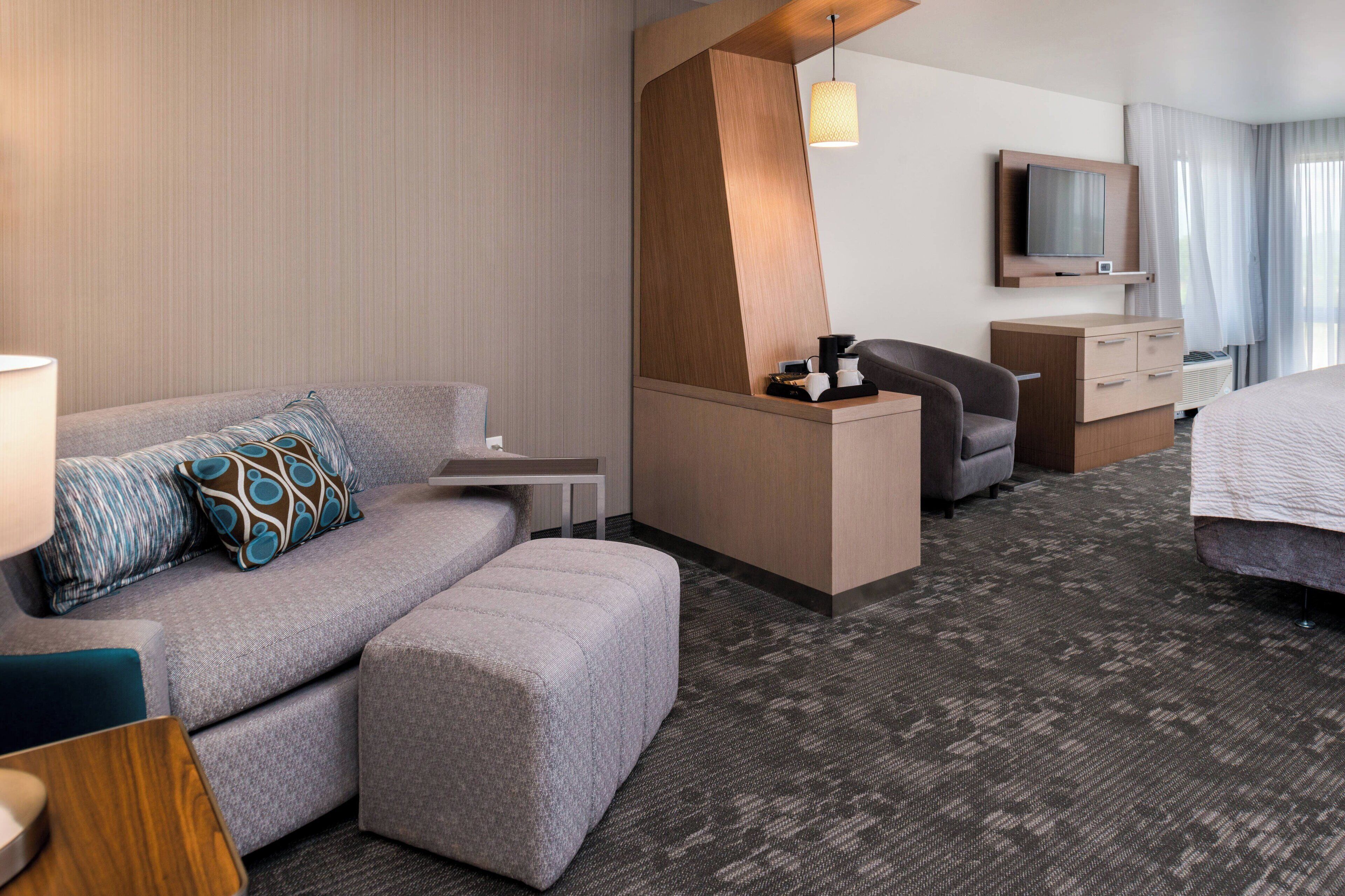 A modern hotel room with a sofa, ottoman, desk, and a TV.