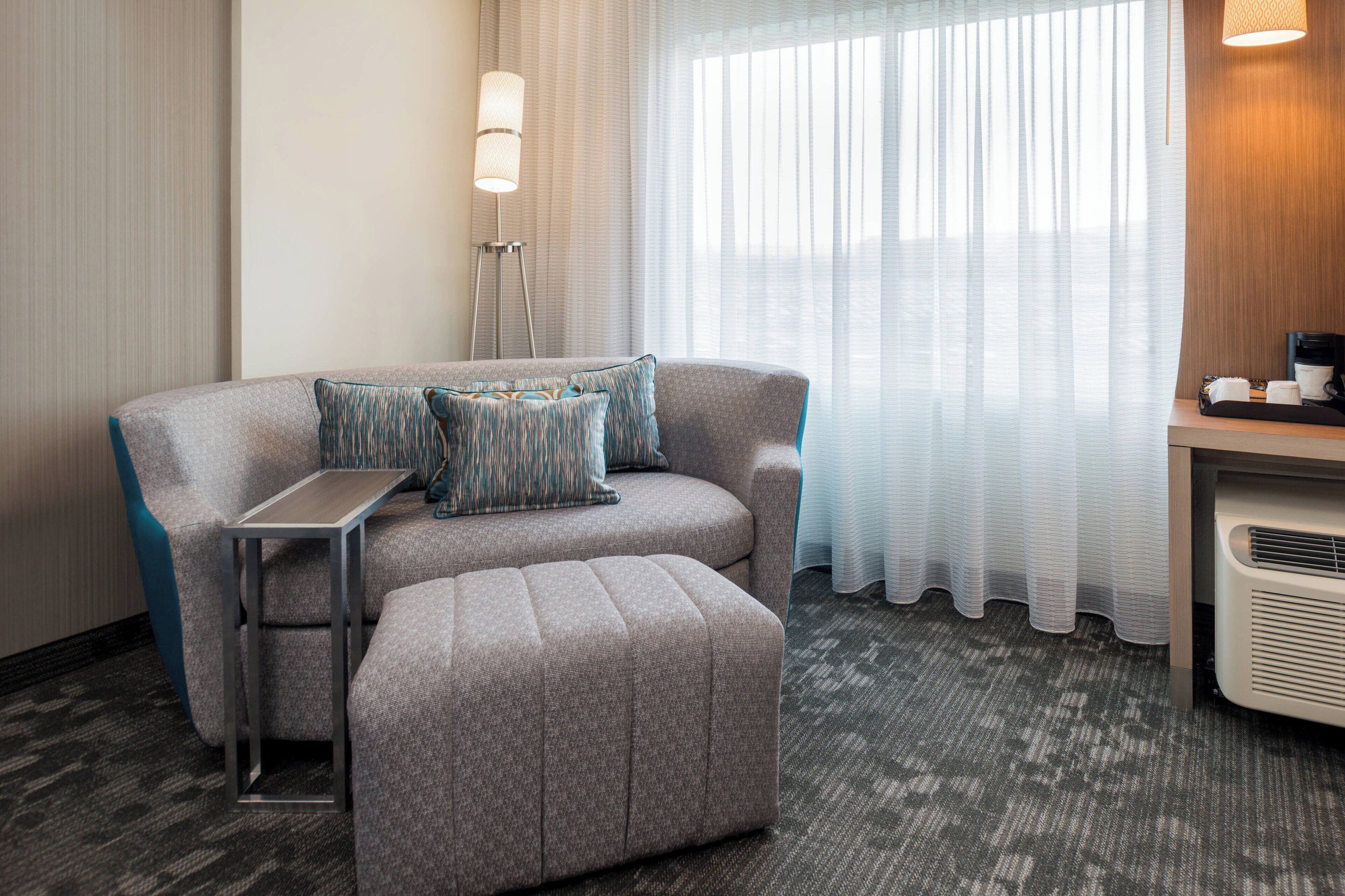 A modern hotel room with a grey sofa, a small table, a standing lamp, and a large window with sheer curtains.