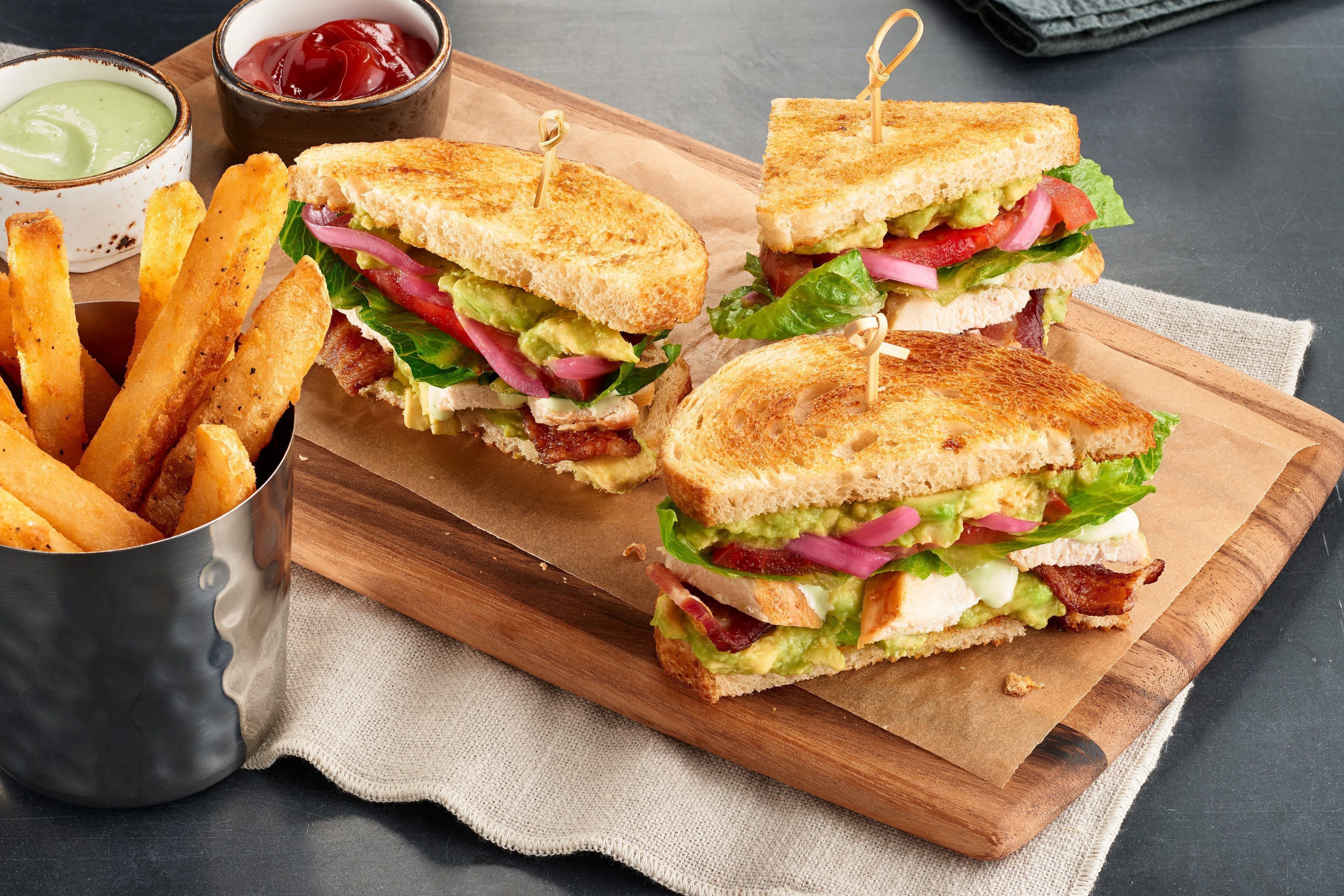 A sandwich with bacon, lettuce, tomato, and avocado on toasted bread, accompanied by French fries and two dipping sauces.
