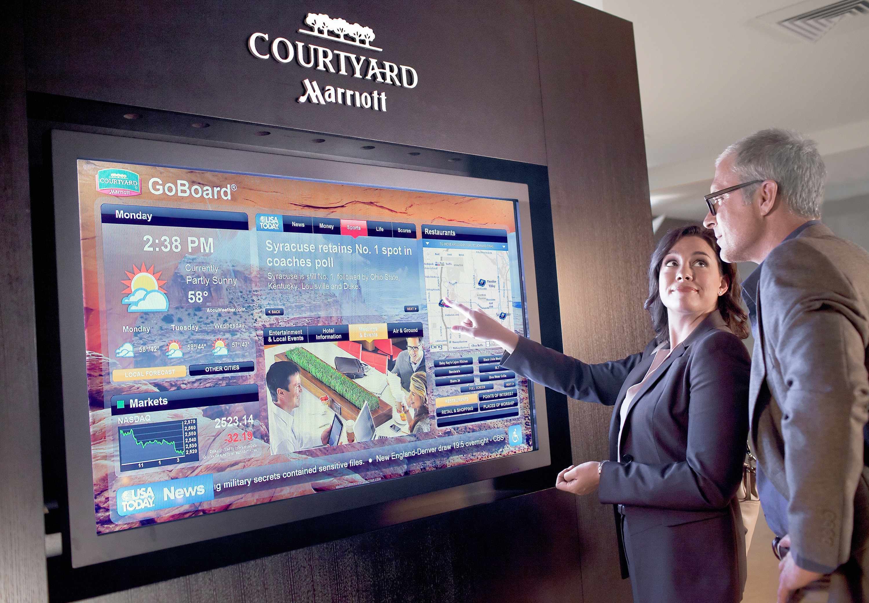 A large digital display at a Courtyard Marriott hotel, featuring a GoBoard interface with local news, weather, and market information.