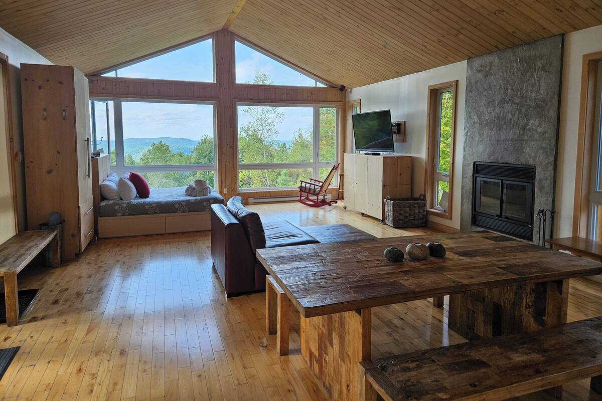 A spacious room with a wooden ceiling, large windows offering a view of the outdoors, a fireplace, and a television mounted on the wall.
