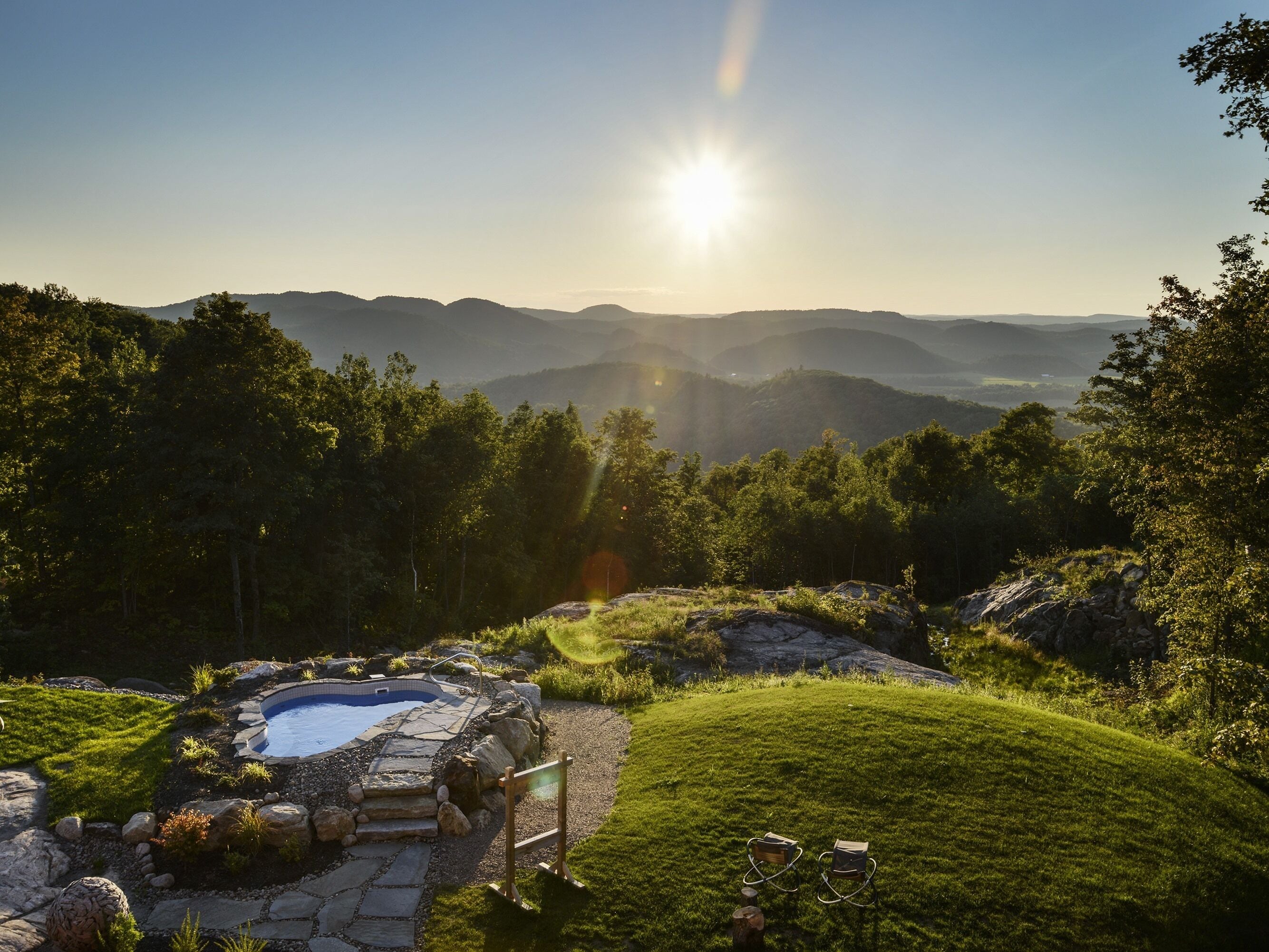 A hot tub surrounded by a stone deck, set in a lush green landscape with a view of distant hills.