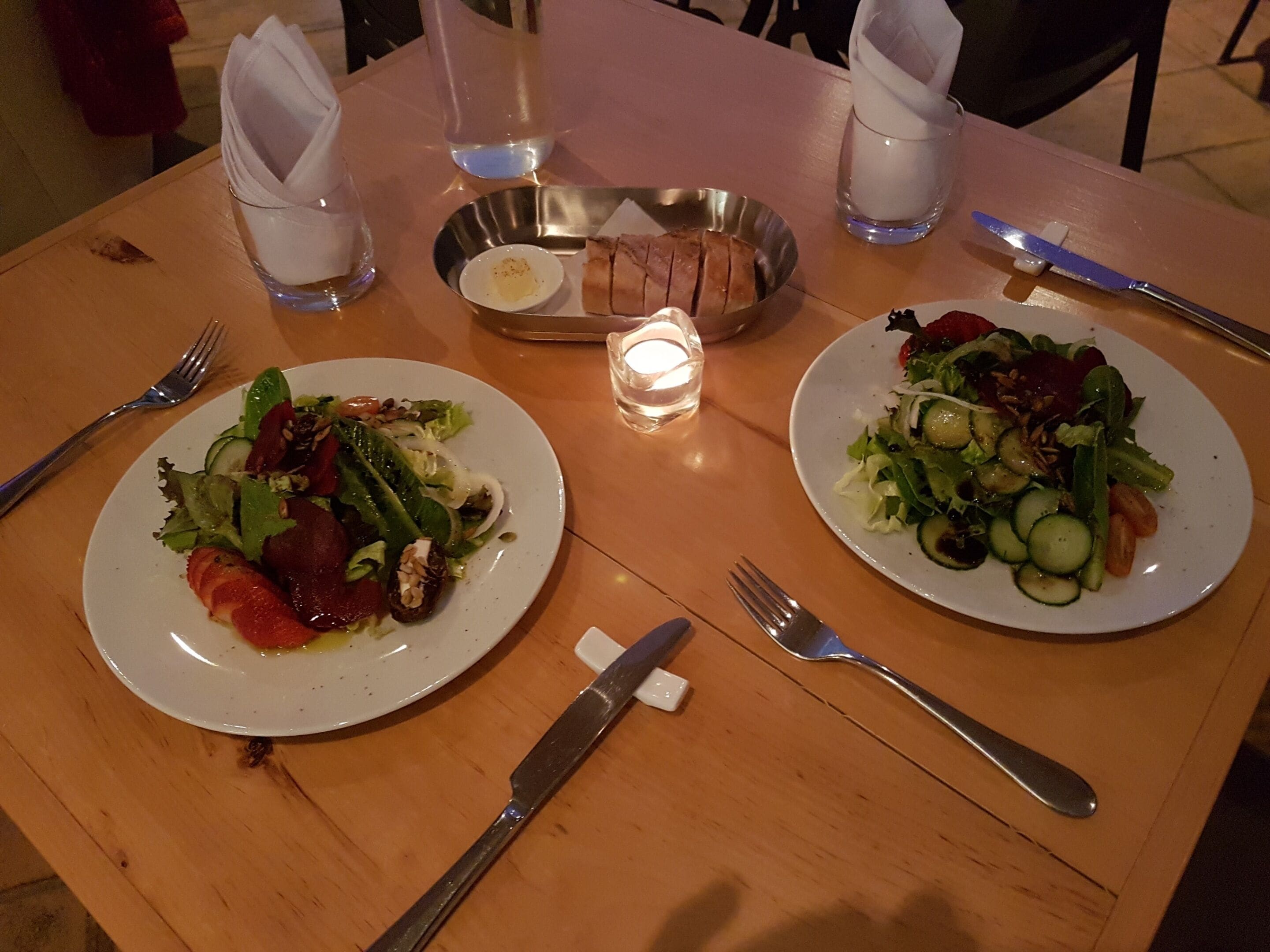 Two plates of food on a wooden table with cutlery and glasses.