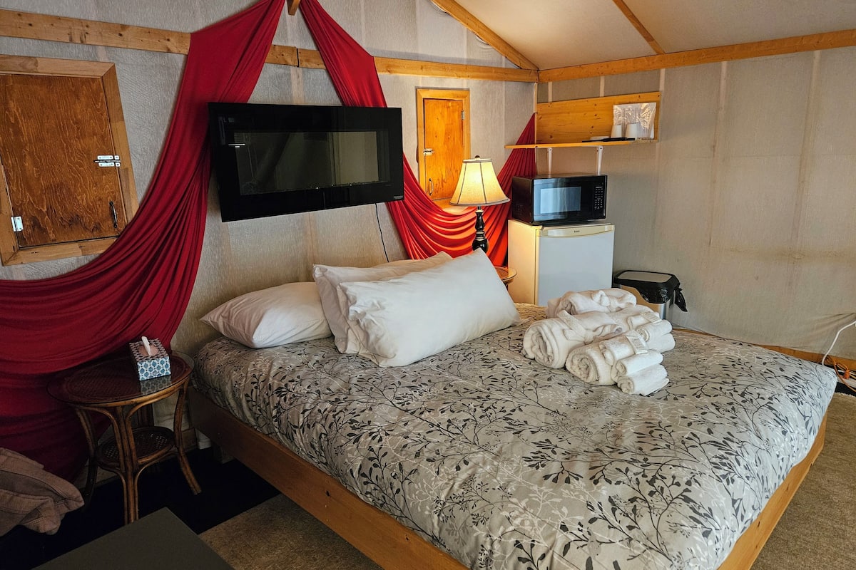 A bedroom with a bed, a TV mounted on the wall, a microwave, and a small table with a lamp.