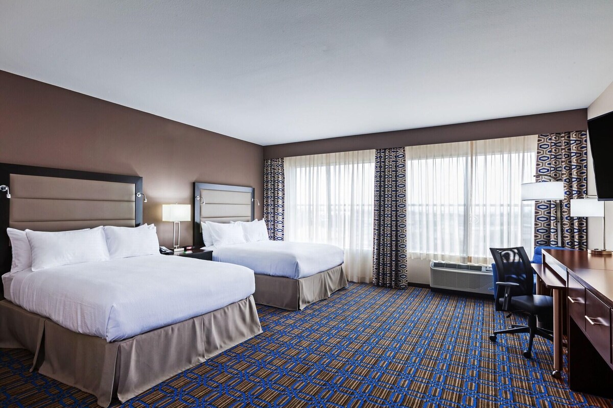 A hotel room with two beds, a desk, a chair, a TV, and a window with patterned curtains.