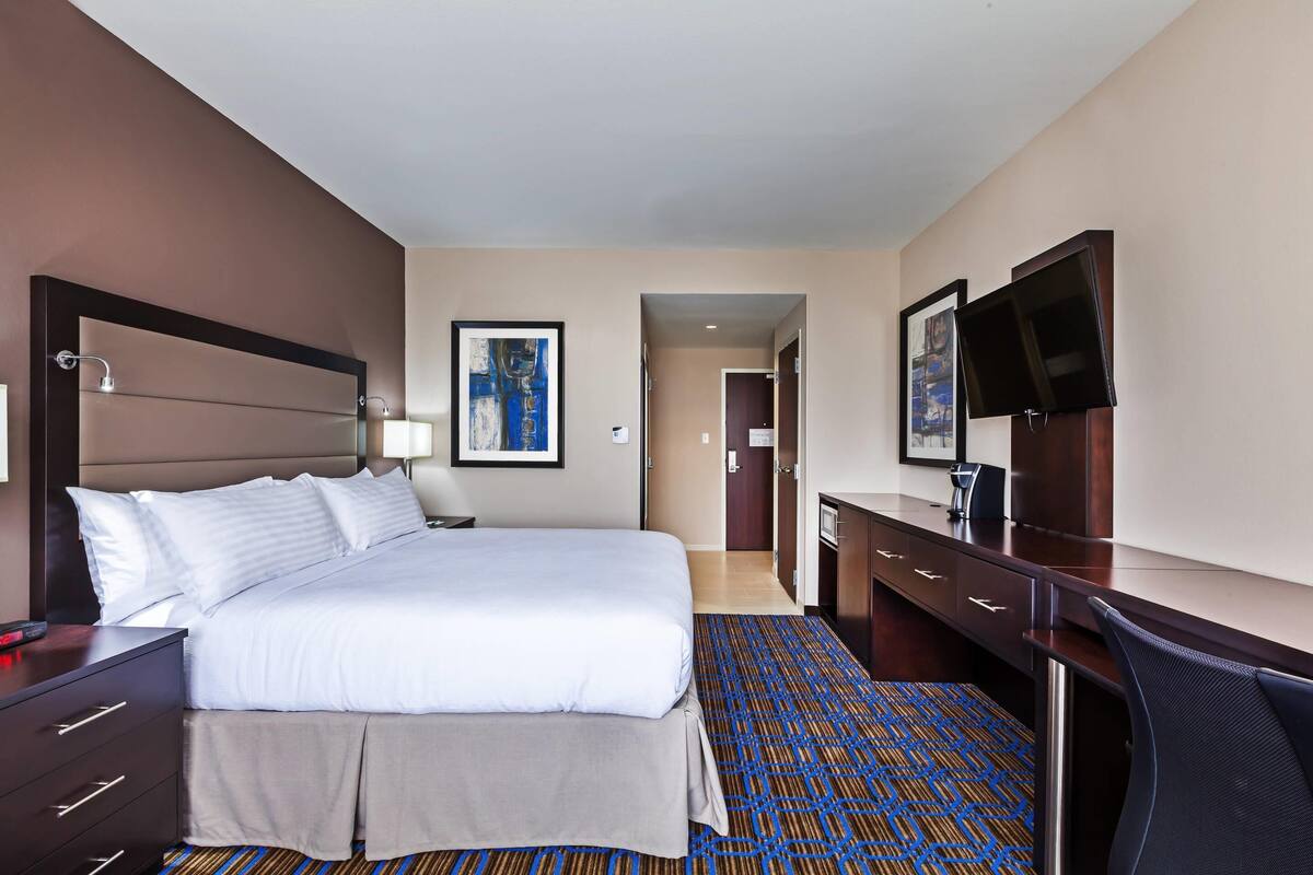 A hotel room with a bed, a desk, a chair, a TV, and a door leading to another room.