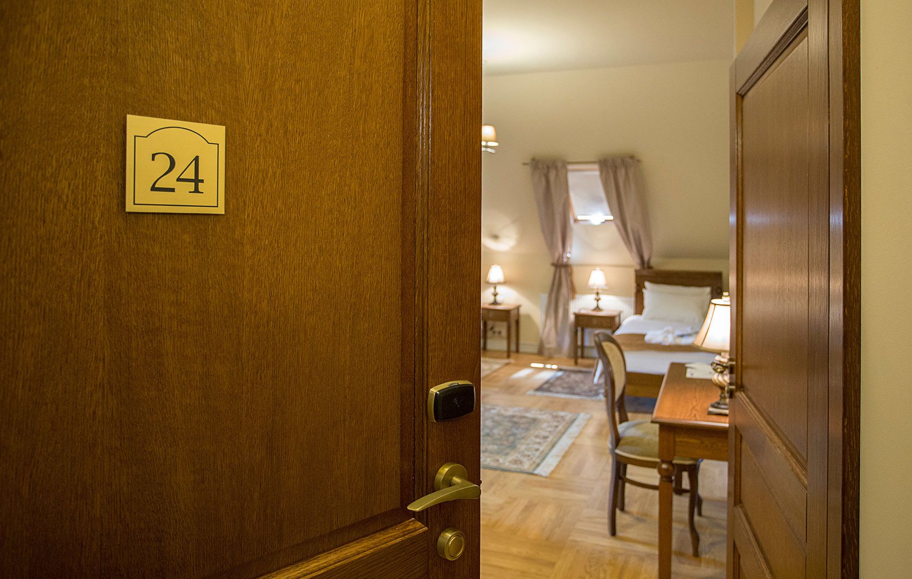 A hotel room with a wooden door labeled "24", a bed with a headboard, two bedside tables with lamps, a chair, and a small table.