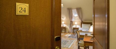 A hotel room with a wooden door labeled "24", a bed with a headboard, two bedside tables with lamps, a chair, and a small table.
