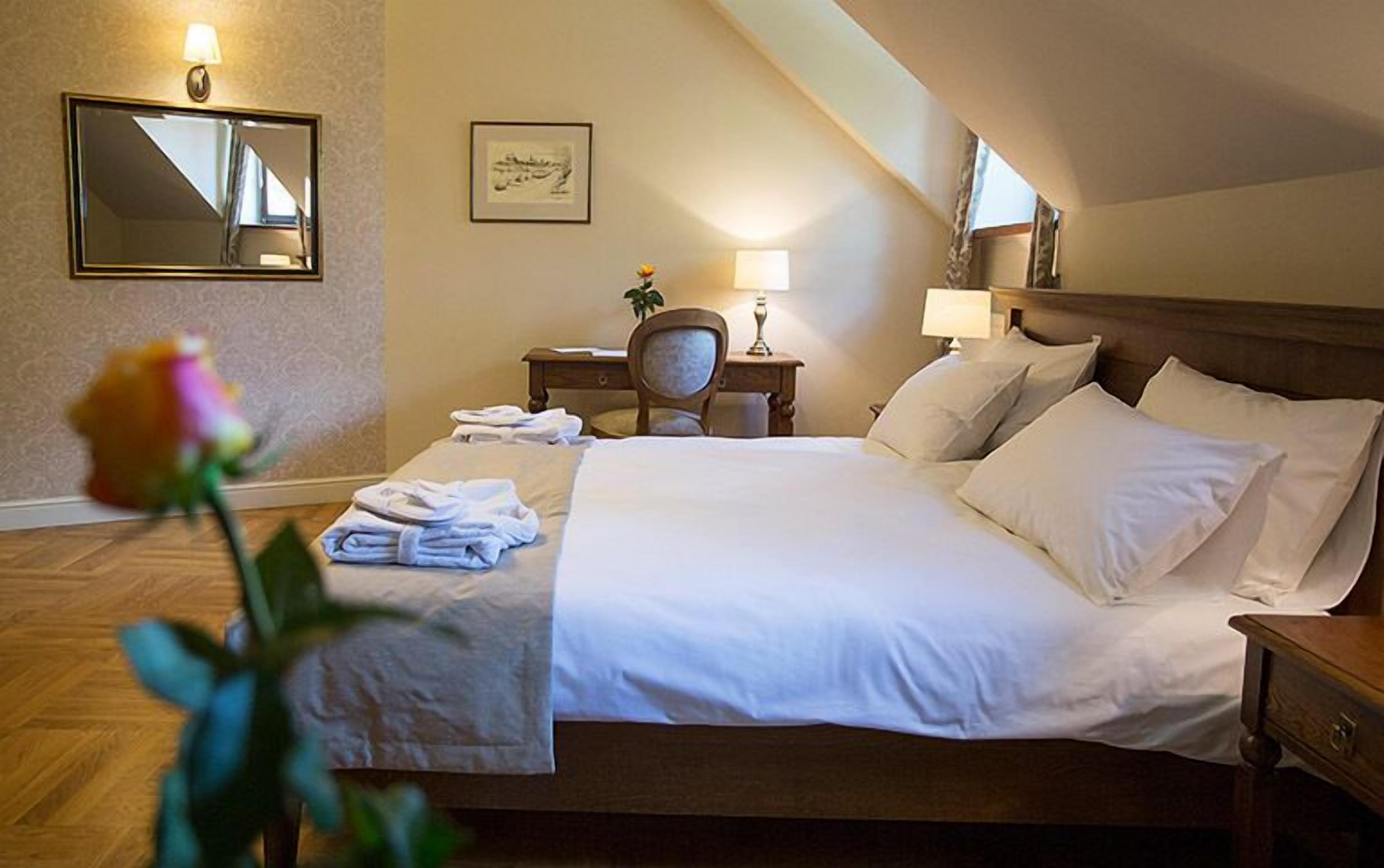 A hotel room with a large bed, a desk, a chair, a nightstand, a lamp, a mirror, and a framed picture on the wall.