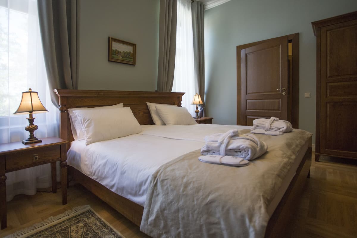 A neatly made bed with white linens and pillows, two bedside lamps, a wooden headboard, a wooden wardrobe, and a small rug on the floor.