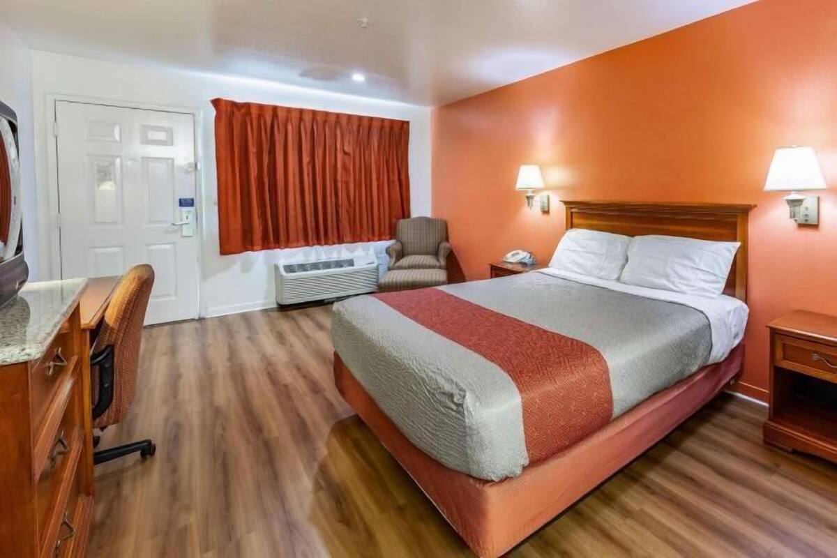 Habitación de hotel con cama, escritorio, silla, mesita de noche y un calentador de pared.