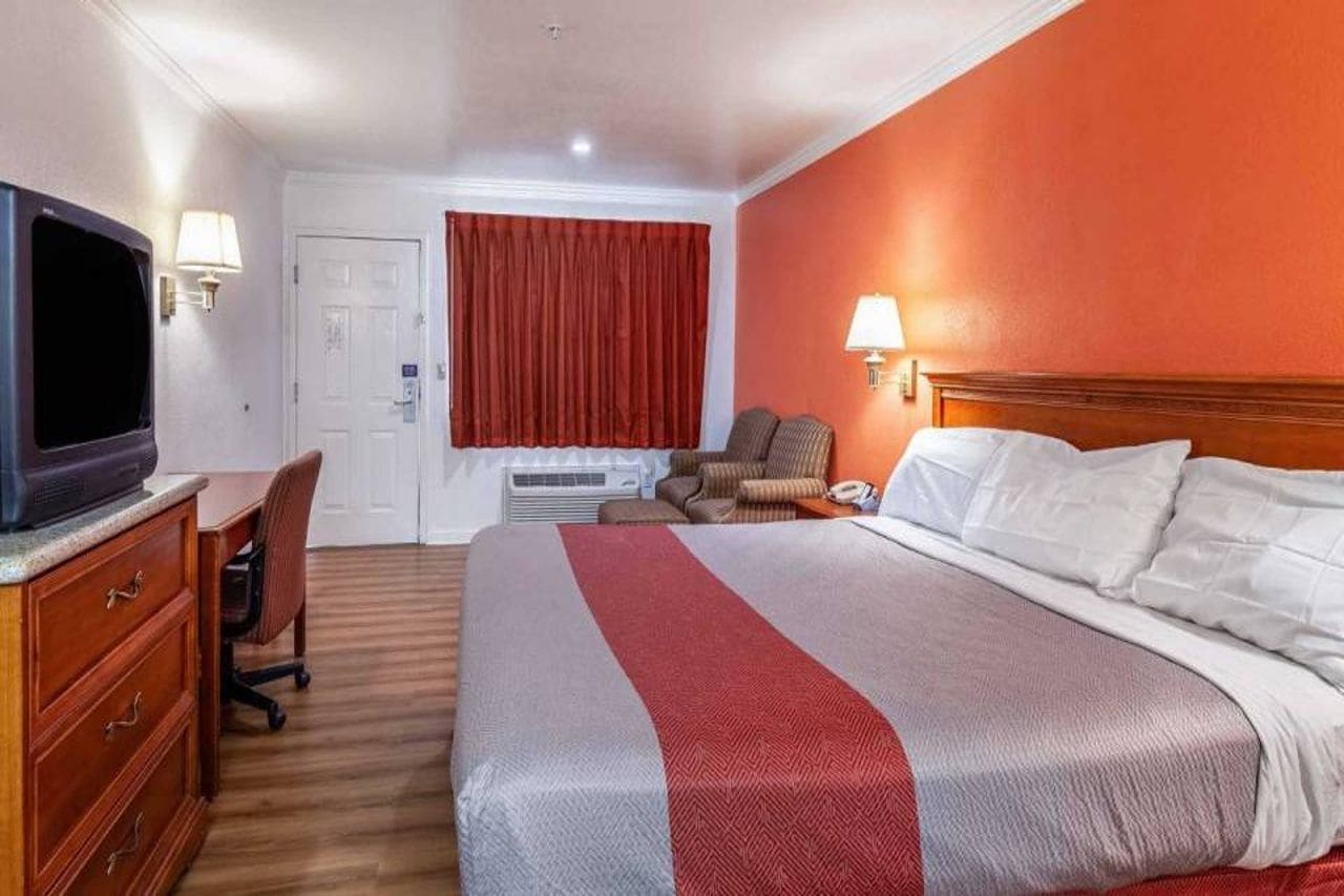 A hotel room with a bed, a desk with a television, a chair, a window with curtains, and a wall-mounted lamp.