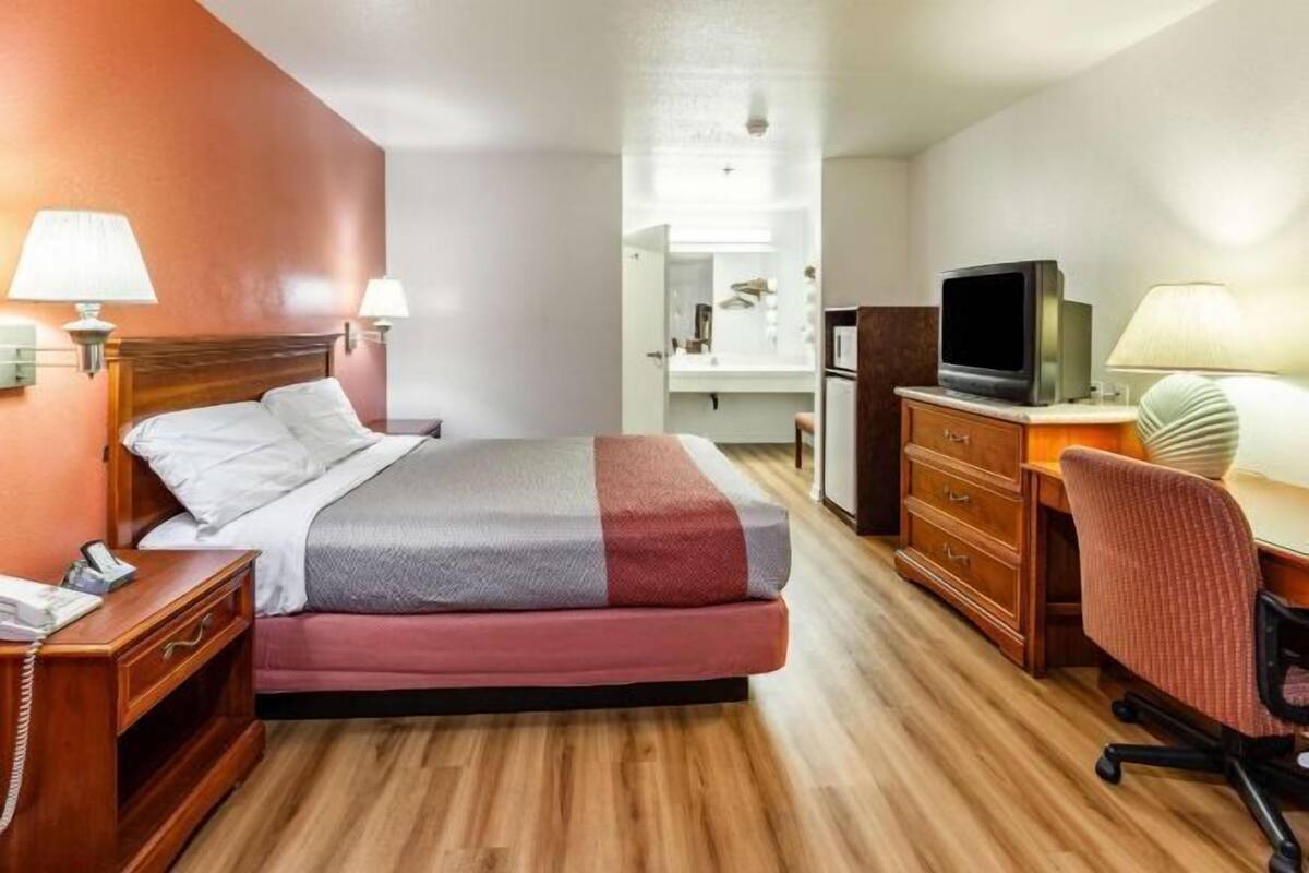 Habitación de hotel con cama, mesita de noche, lámpara, televisor, escritorio, silla y una pequeña cocina.