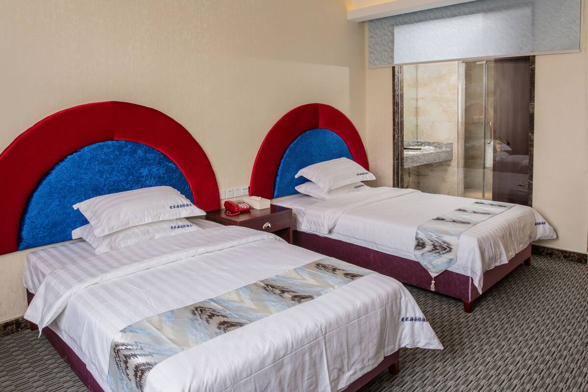 Habitación de hotel con dos camas, cada una con cabecero rojo y azul, ropa de cama blanca y una mesita de noche pequeña con un teléfono rojo.