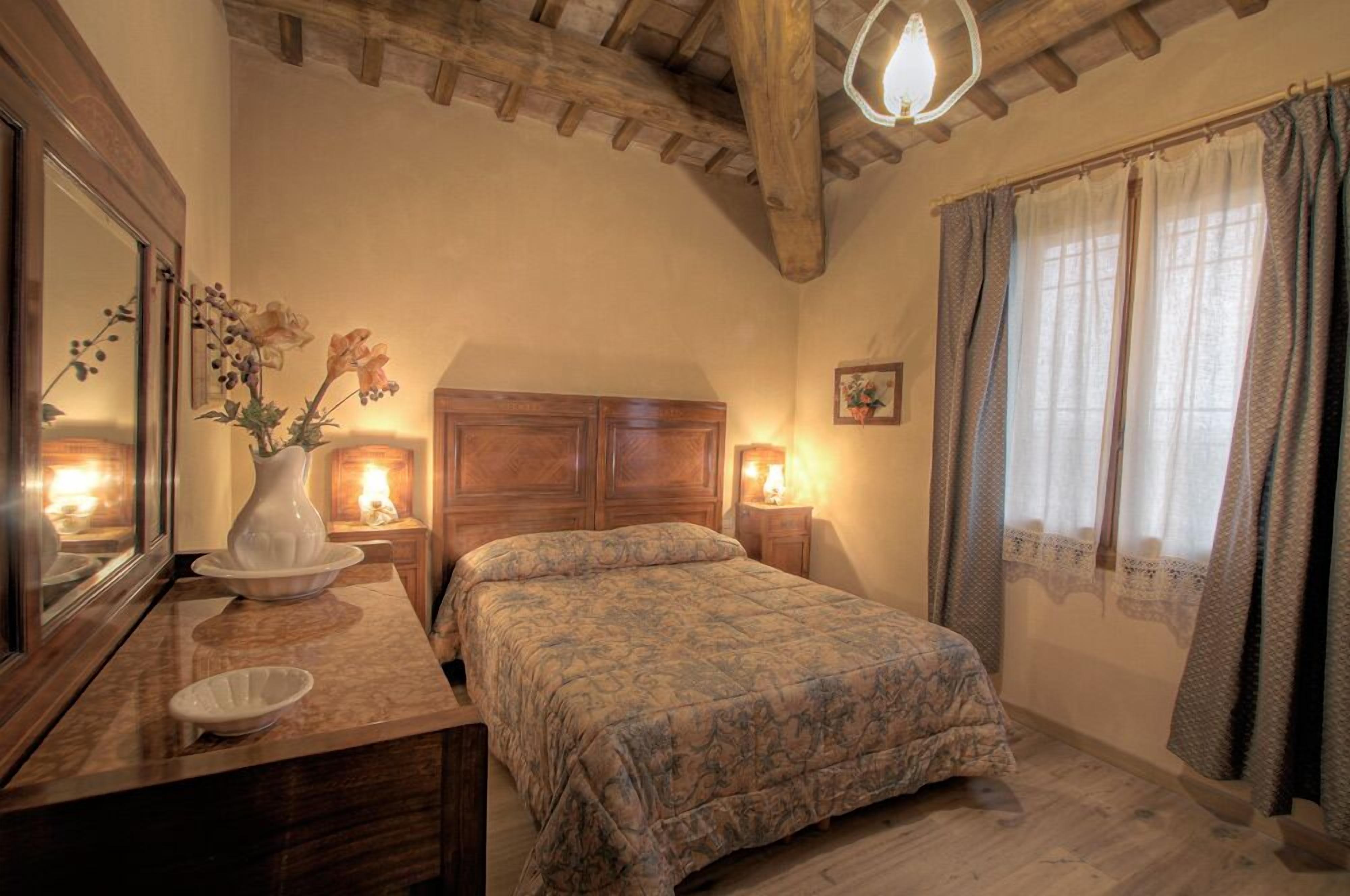 Un dormitorio con cabecera de madera, cama con colcha estampada, mesita de noche con lámpara, espejo y un florero con flores.