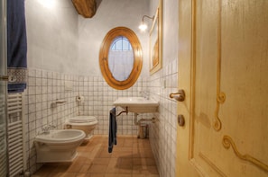 A bathroom with a toilet, bidet, and sink, a wooden door, and a window with a wooden frame. - La Fornasaccia (Cesena)
