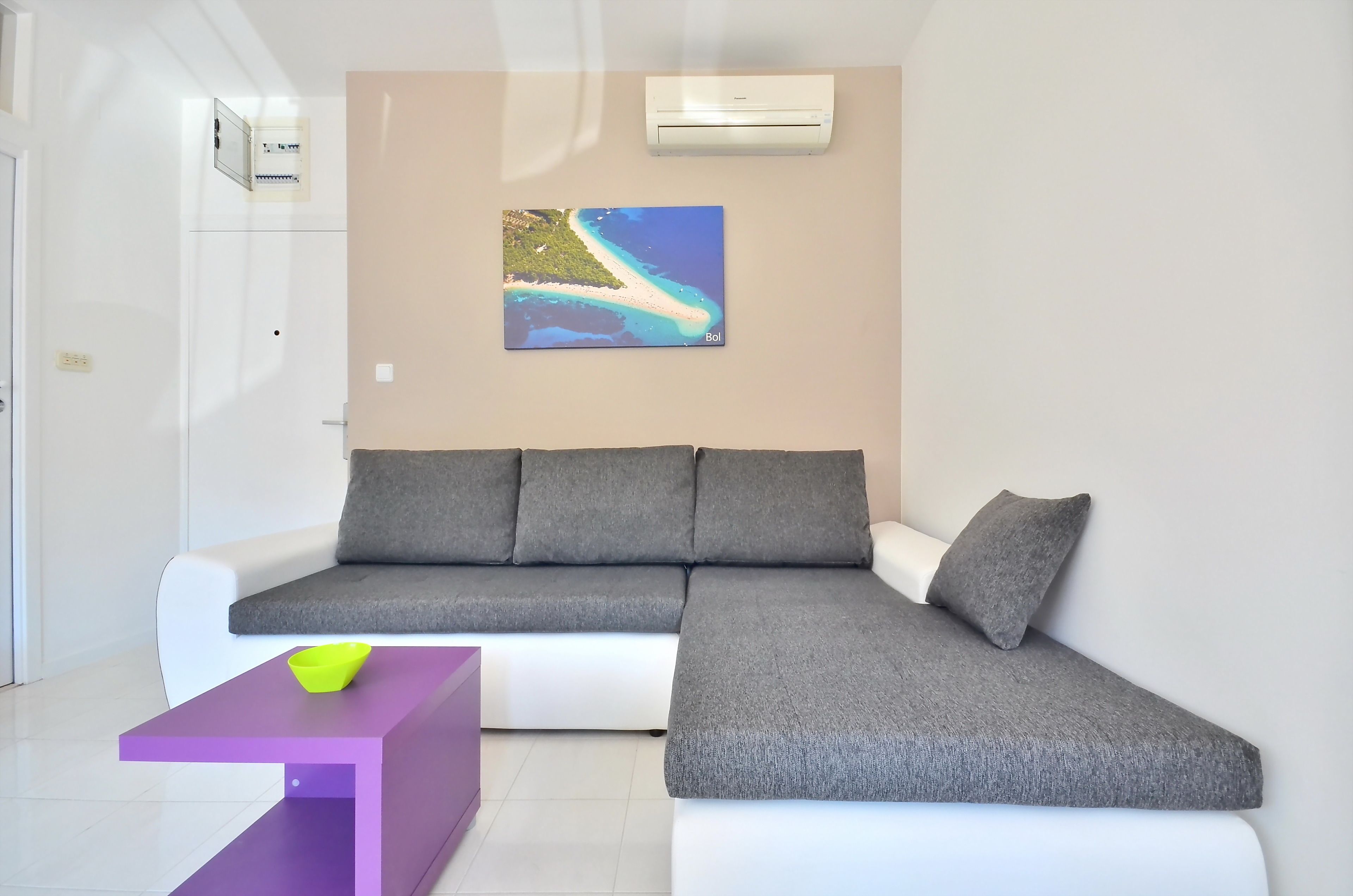 Ein modernes Wohnzimmer mit einem grauen Ecksofa, einem violetten Beistelltisch und einer Wand-Klimaanlage.