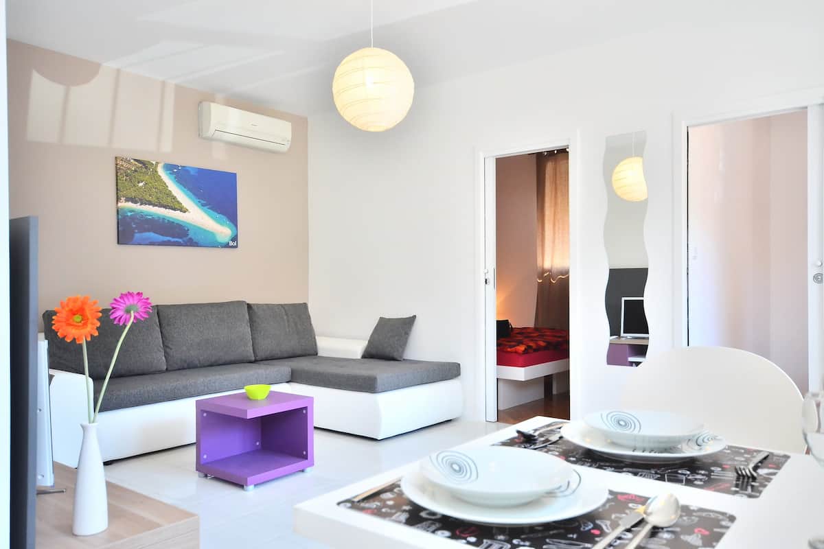 Una sala moderna con un sofá gris, una mesita auxiliar morada y un comedor con una mesa blanca dispuesta para dos personas.