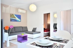 Una sala moderna con un sofá gris, una mesita auxiliar morada y un comedor con una mesa blanca dispuesta para dos personas.