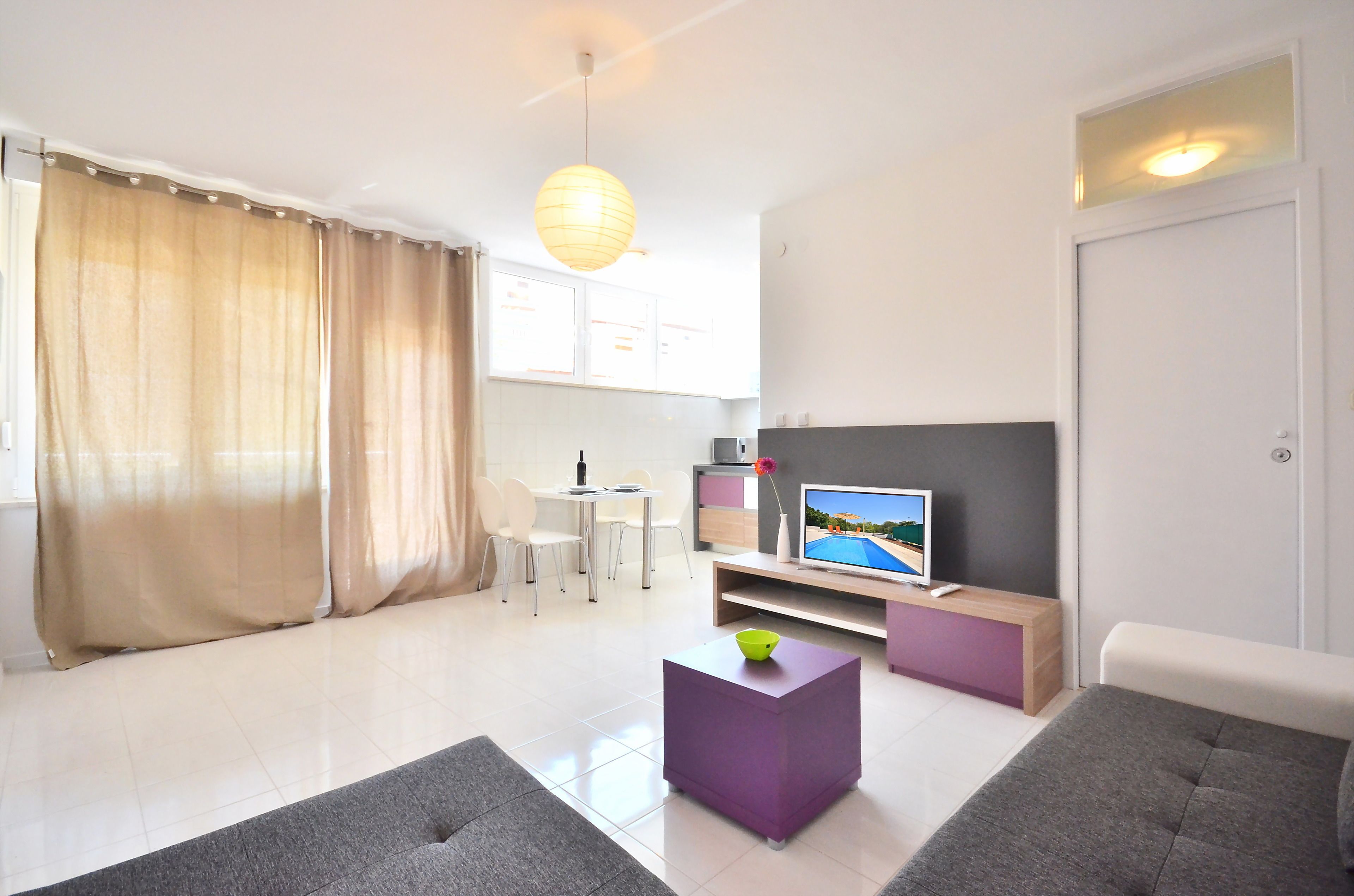 A modern living room with a flat-screen TV, a dining area with chairs, a kitchen area, and a large window with curtains.
