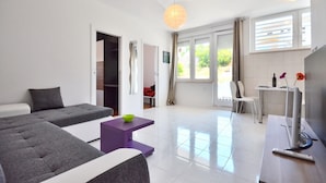 A modern living room with a sofa, a small table, and a TV. There is a door leading to another room and a window with curtains. - BRETIA apartments & more (Sutivan)