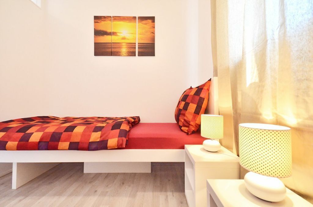 Een moderne slaapkamer met een bed, een nachtkastje met een lamp en een aan de muur hangend schilderij van een zonsondergang.