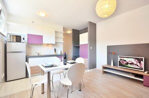 A modern kitchen with white cabinets, a dining table with chairs, a TV on a stand, and a refrigerator. - BRETIA apartments & more (Sutivan)