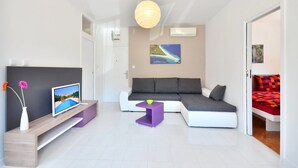 A modern living room with a sectional sofa, a flat-screen TV, a small purple side table, and a vase with flowers. - BRETIA apartments & more (Sutivan)