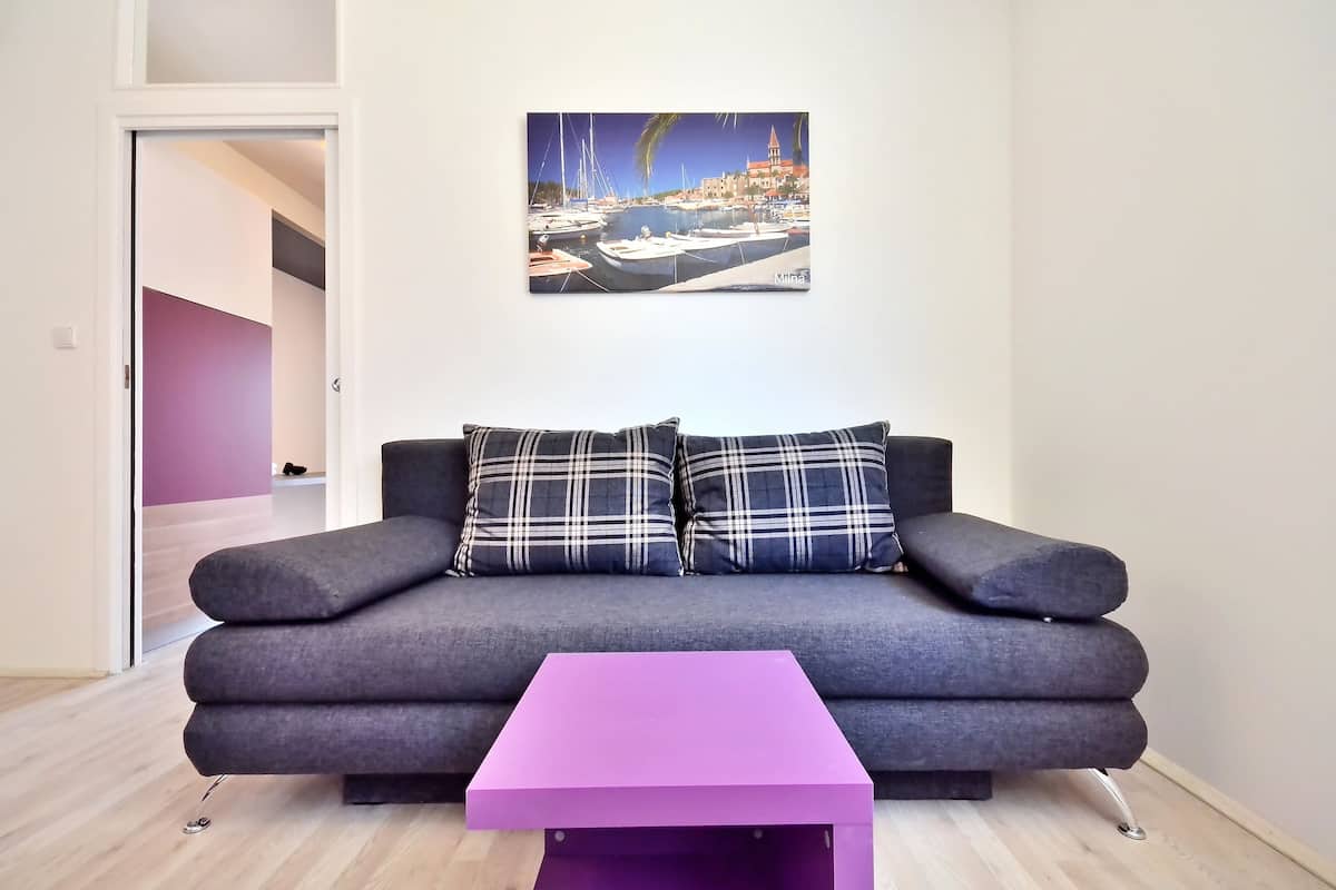 Una sala moderna con un sofá gris, dos cojines a cuadros, una mesa de centro morada y un cuadro con una escena costera en la pared.