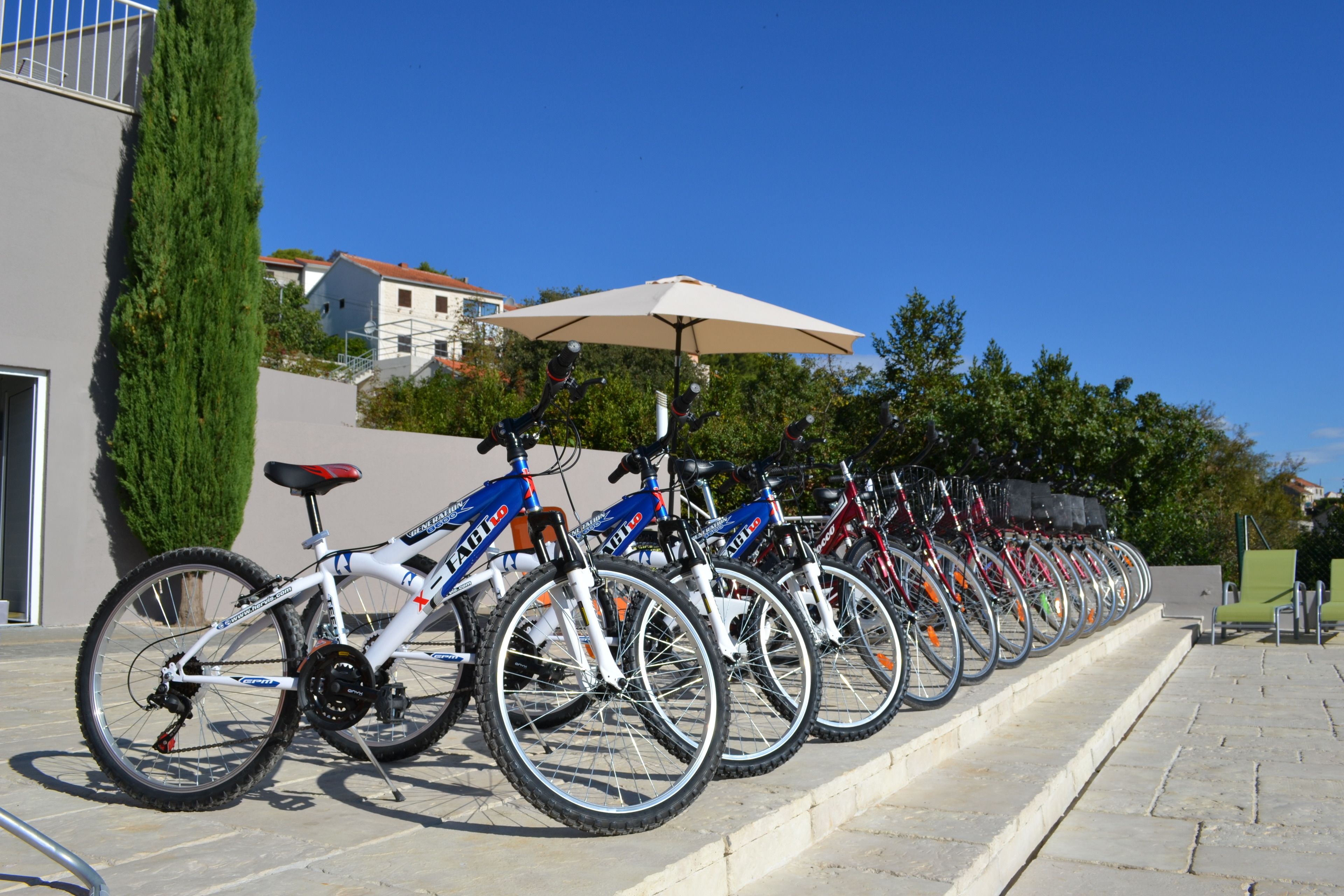 A row of bicycles parked on a stone pavement, with a large umbrella providing shade.