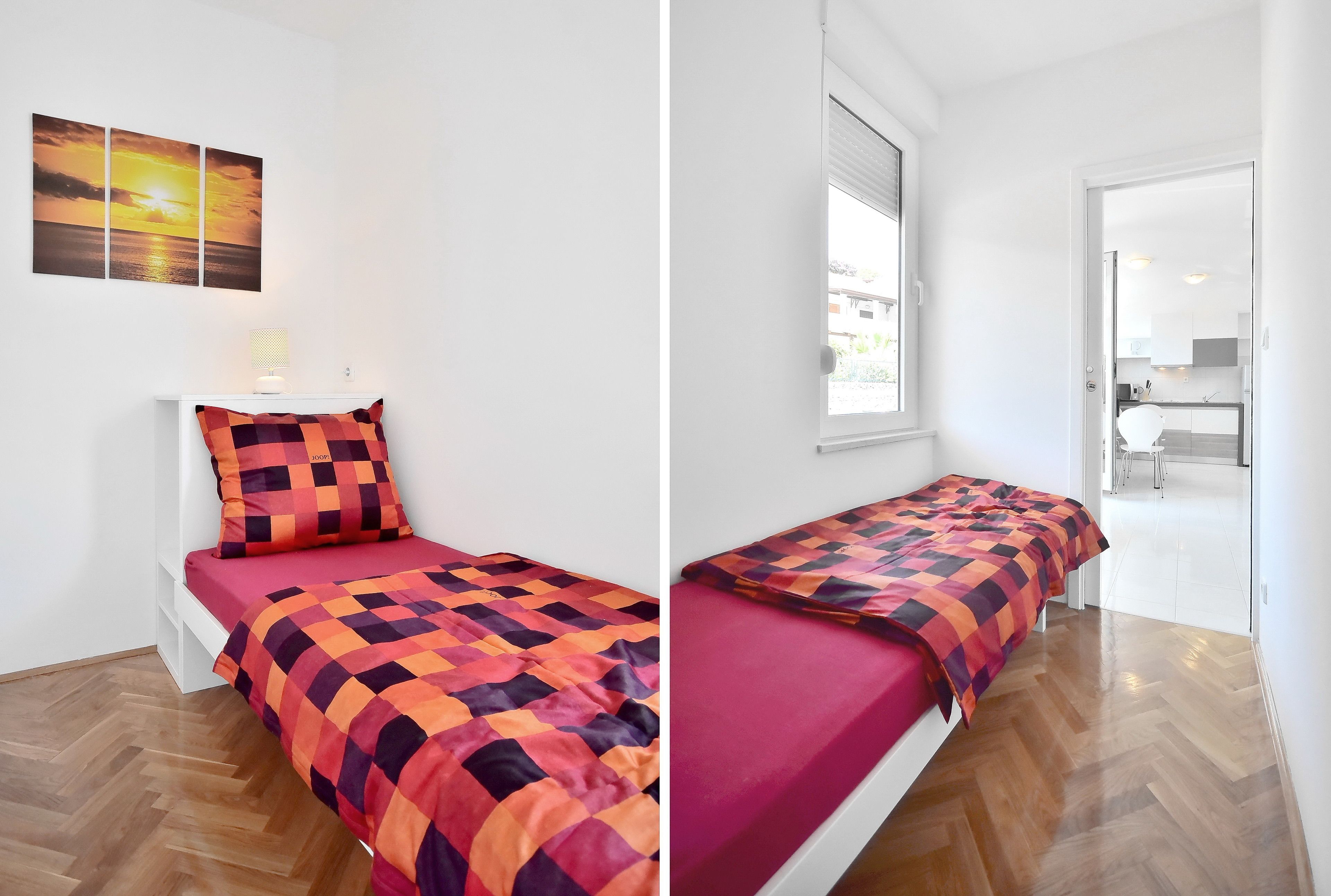 Una cama individual con colcha a cuadros de colores vivos y almohada a juego, una mesita de noche blanca con lámpara, y un cuadro colgado en la pared que muestra un atardecer.