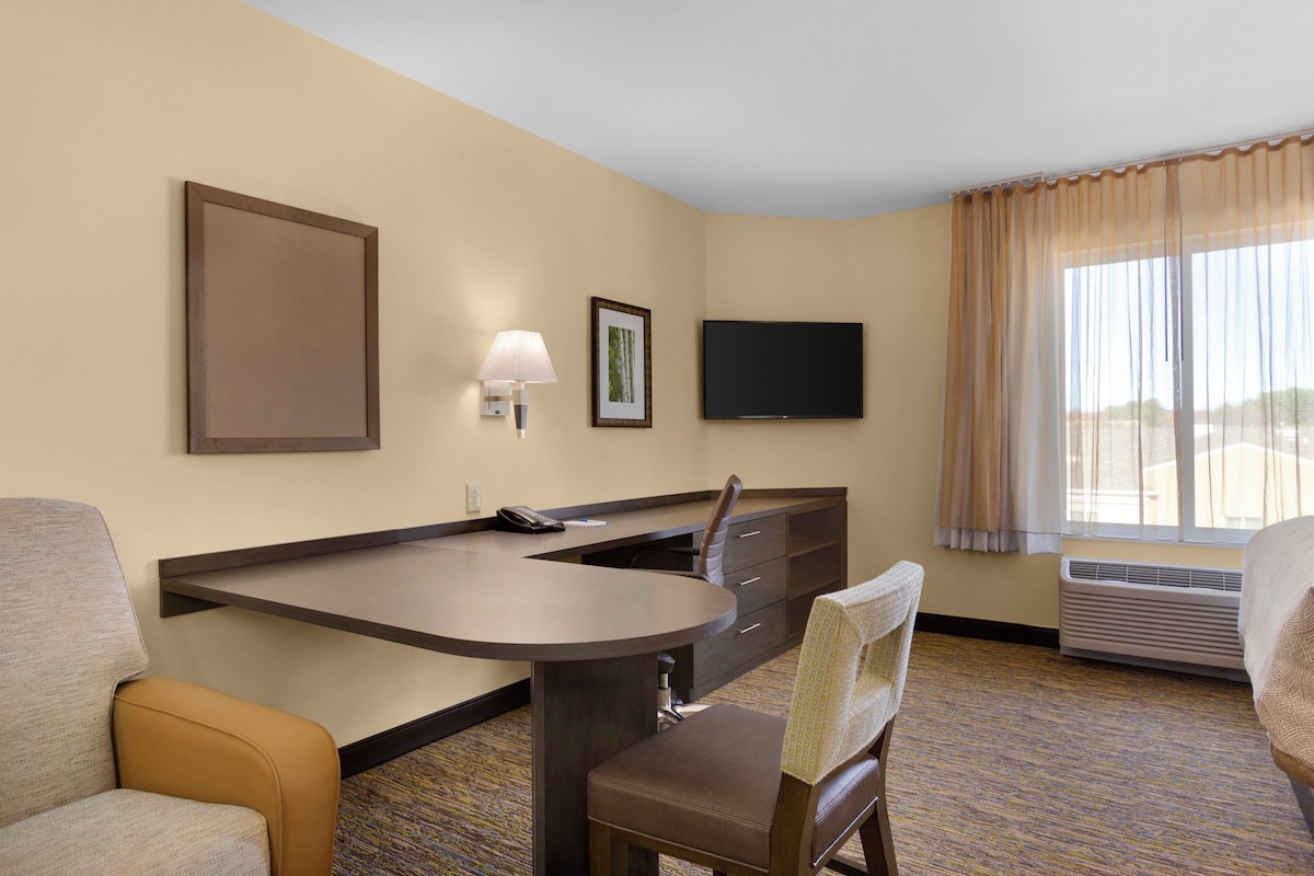 A hotel room with a desk, two chairs, a flat-screen TV, a framed picture, and a window with curtains.