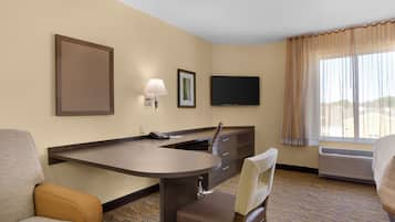 A hotel room with a desk, two chairs, a flat-screen TV, a framed picture, and a window with curtains.