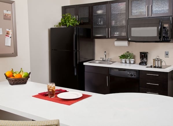 A modern kitchen with dark cabinets, a black refrigerator, a microwave, and a sink. There is a fruit basket and a glass of iced tea on the counter. - Candlewood Suites Vestal - Binghamton by IHG (Vestal)