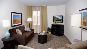 A hotel room with a brown sofa, a small round table, a TV, a lamp, and a window with curtains. - Candlewood Suites Vestal - Binghamton by IHG (Vestal)