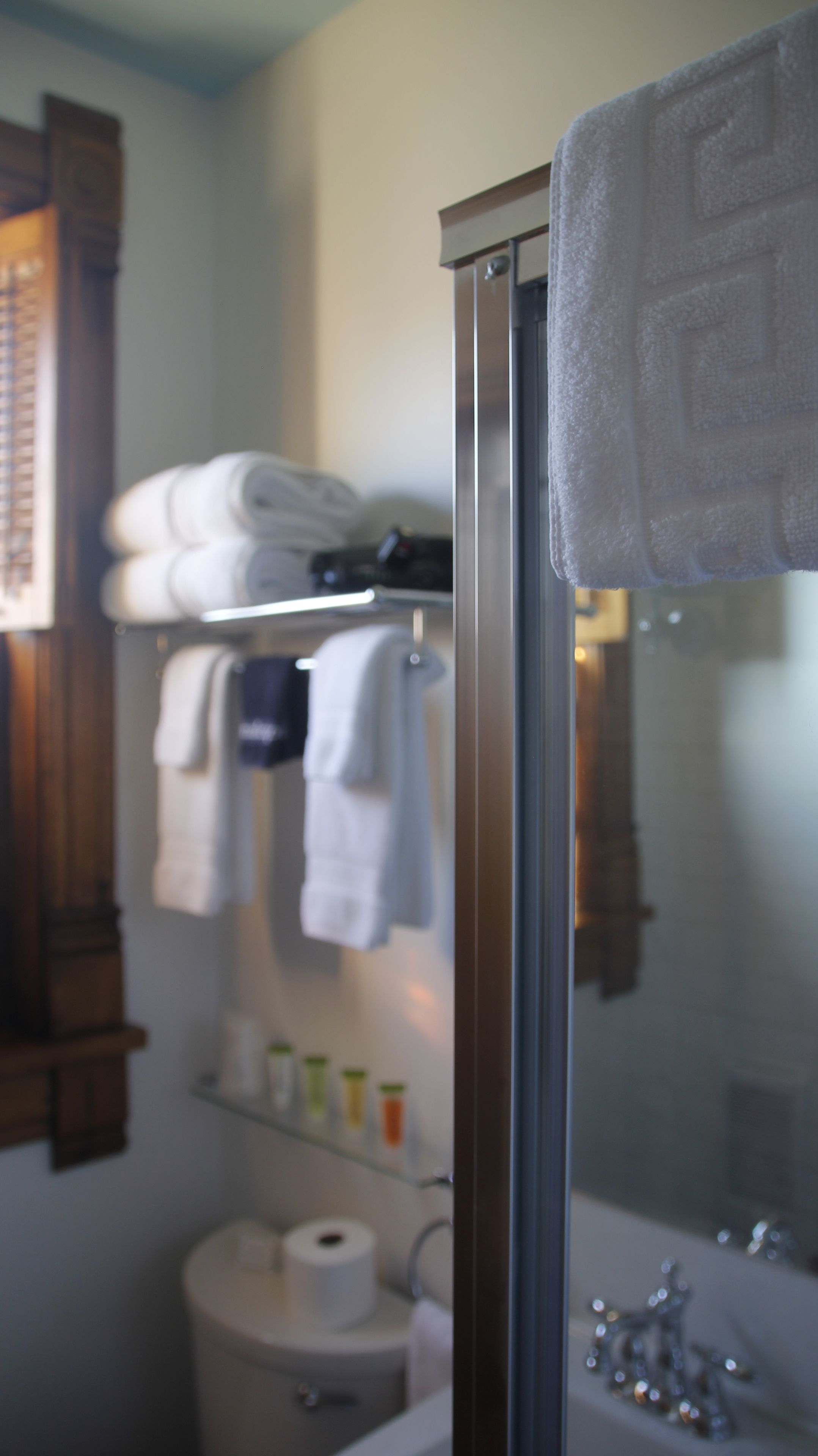 A bathroom with a glass shower enclosure, a towel rack with folded towels, a shelf with toiletries, and a toilet.