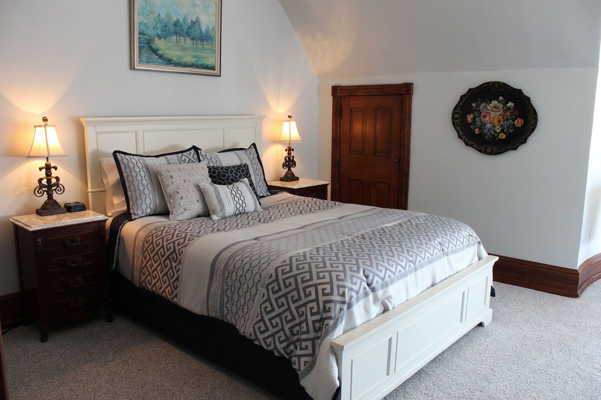 Une chambre à coucher bien rangée, avec un lit, deux tables de chevet, une petite table, une porte en bois et une assiette décorative accrochée au mur.