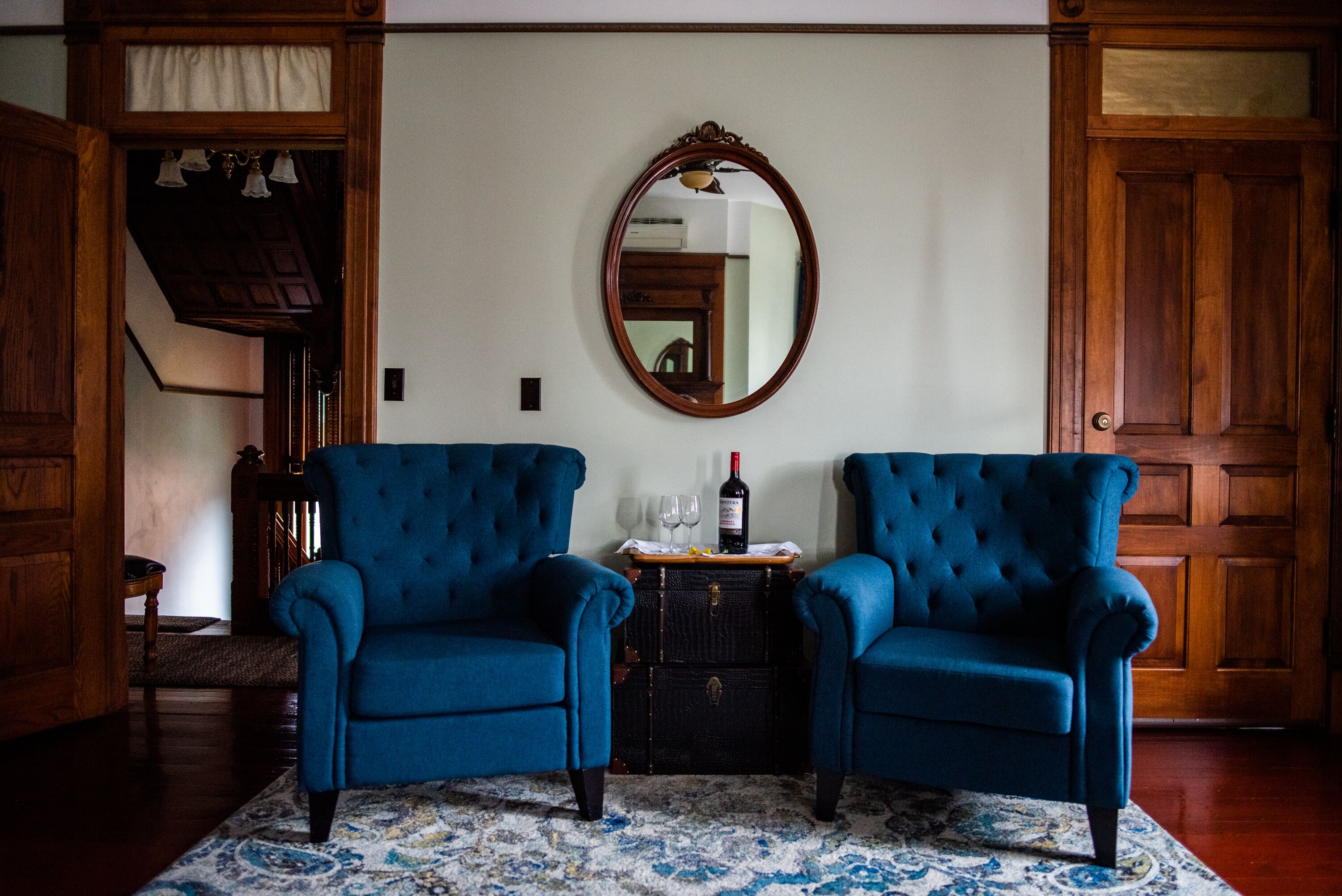 A room with two blue armchairs, a wooden chest of drawers, a mirror, and a bottle of wine on the chest.