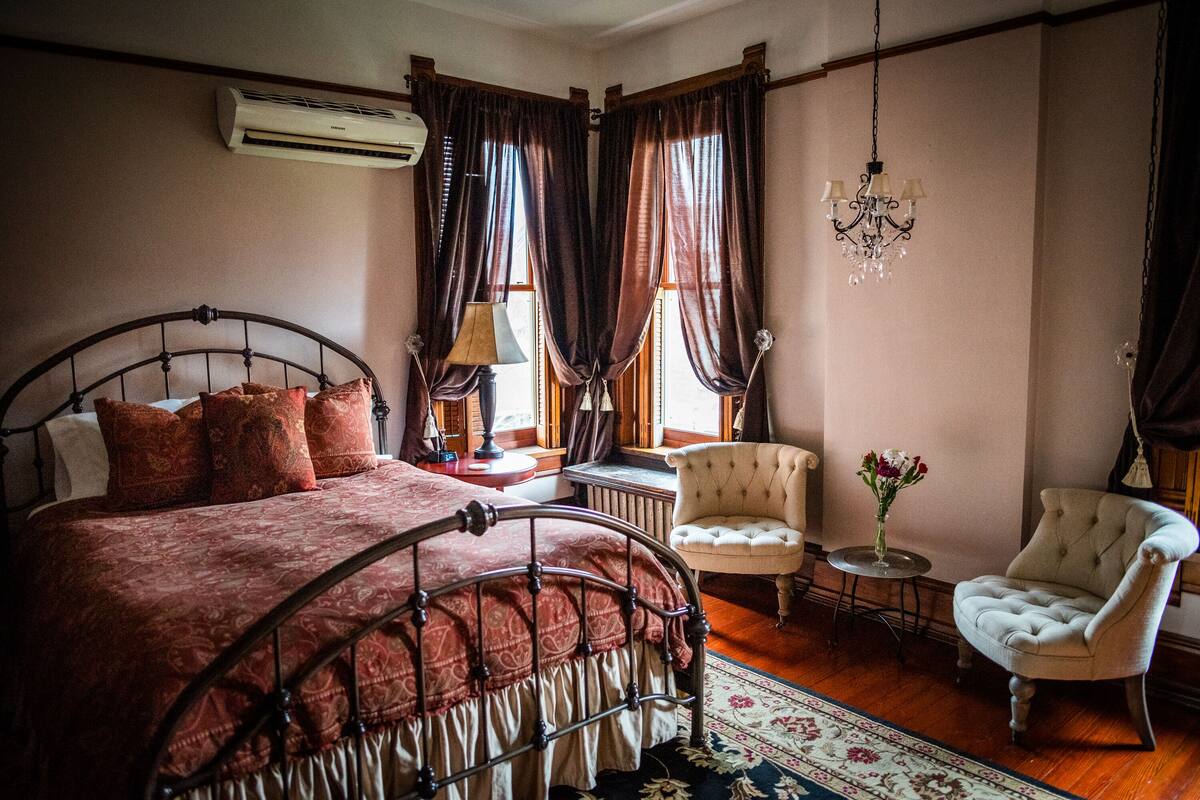Une chambre à coucher avec un lit en métal, une couvre-lit rouge, un lustre, deux chaises, une petite table avec une lampe et un vase de fleurs.