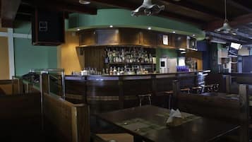 Zona de bar con asientos de madera, mesas y una barra con botellas, además de una lámpara colgante.