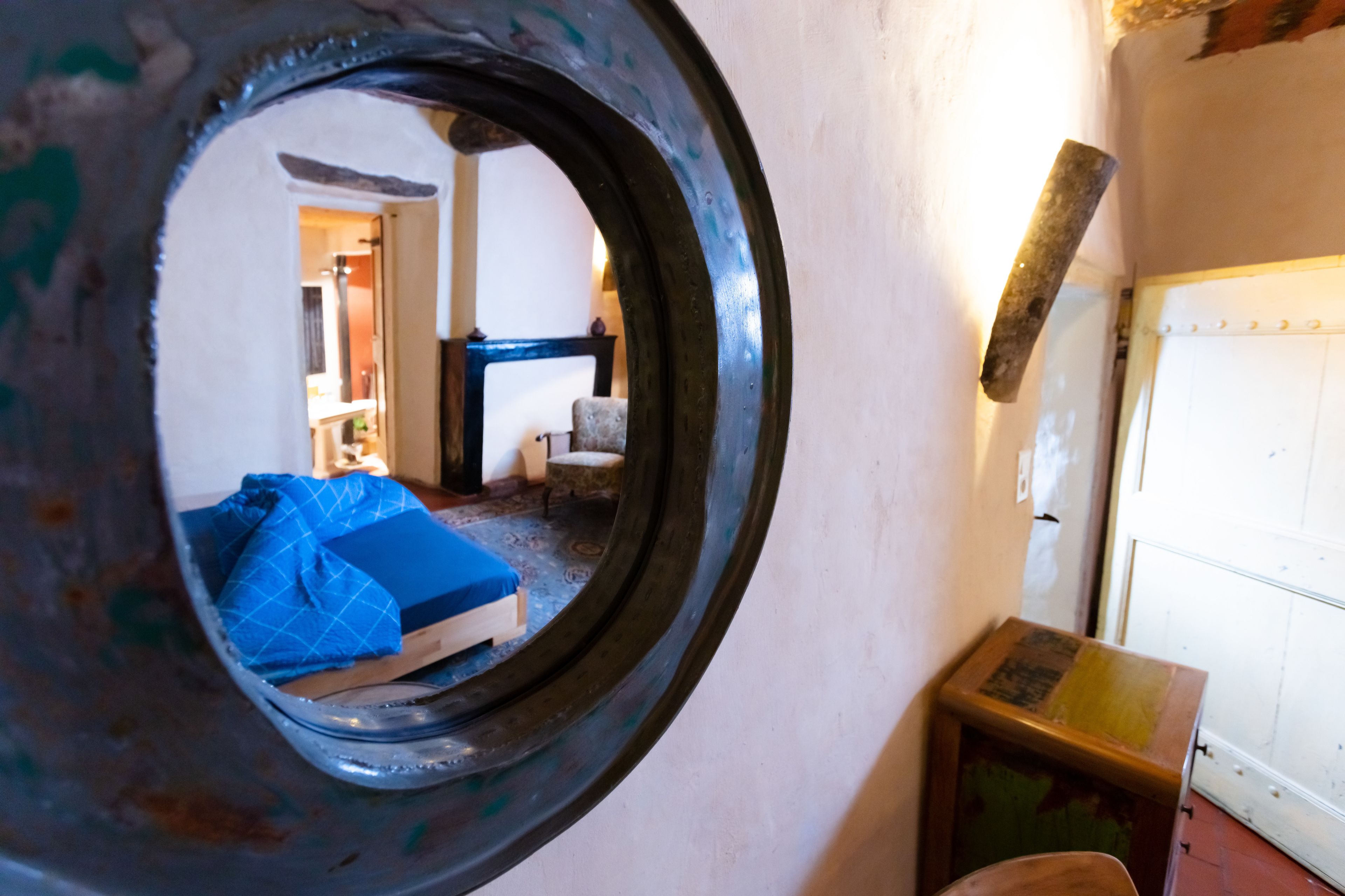 A room with a circular window, a bed with blue bedding, a small table, and a radiator.