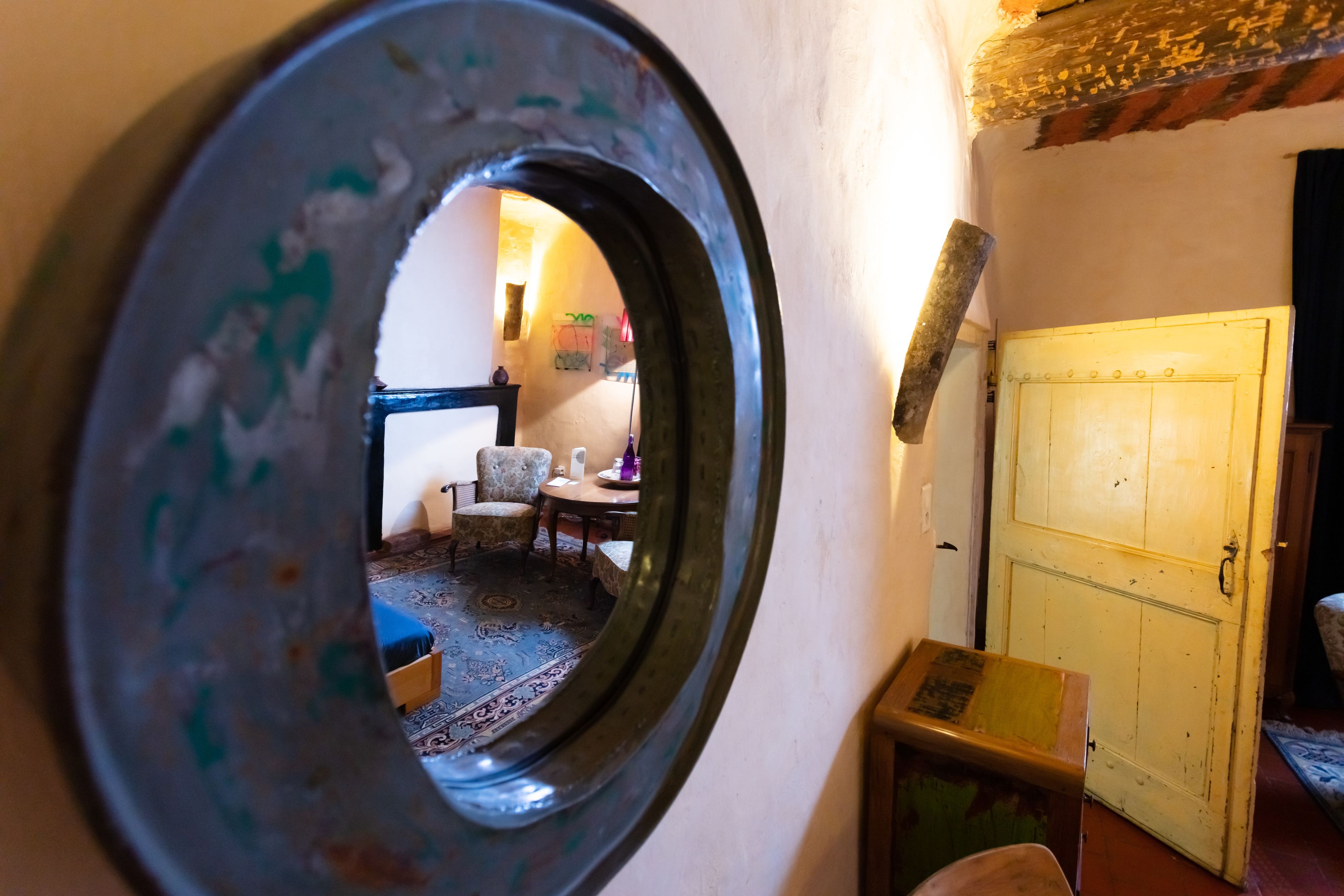 A round mirror reflecting a room with a patterned rug, a wooden chair, and a yellow door.